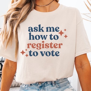 Op de afbeelding: Een crèmekleurig T-shirt met korte mouwen en de tekst "ask me how to register to vote" in donkerblauw en terracotta op de voorzijde. Het ontwerp bevat kleine vierpuntige sterren in terracotta die de tekst omringen.