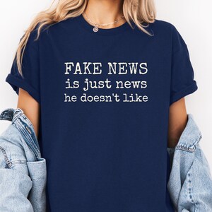 May include: A navy blue short-sleeved t-shirt featuring white text printed on the chest that reads: FAKE NEWS is just news he doesn't like. The shirt is worn with a light blue denim jacket draped over the shoulders, showcasing a casual style.