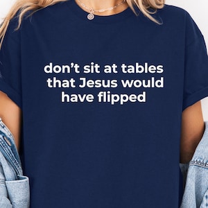 Peut inclure: T-shirt bleu marine avec le texte blanc : « ne vous asseyez pas aux tables que Jésus aurait renversées ». Le t-shirt a des manches courtes et un col rond. Le t-shirt est fait d'une matière douce.