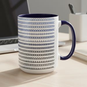 May include: White ceramic mug with a navy blue interior and handle. The mug is decorated with rows of blue and black line art patterns. The mug is sitting on a light wood surface.