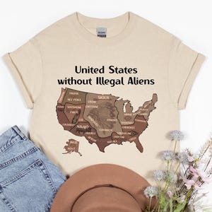 May include: Beige t-shirt with the text "United States without Illegal Aliens" above a map of the United States. The map is brown and features the names of Native American tribes. A pair of blue jeans and a brown hat are in the image.