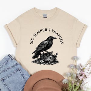 May include: A beige t-shirt featuring a black illustration of a raven perched atop a crown. The text "SIC SEMPER TYRANNIS" is arched above the raven. The shirt is folded, with a pair of blue jeans and a brown hat in the background.