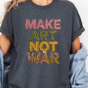 K&ouml;nnte beinhalten: Dunkelgraues T-Shirt mit dem Schriftzug "MAKE ART NOT WAR" in strukturierten, farbenfrohen Buchstaben. Der Text ist in Rosa, Gr&uuml;n, Gelb und Rosa gehalten. Das T-Shirt hat hochgekrempelte &Auml;rmel.