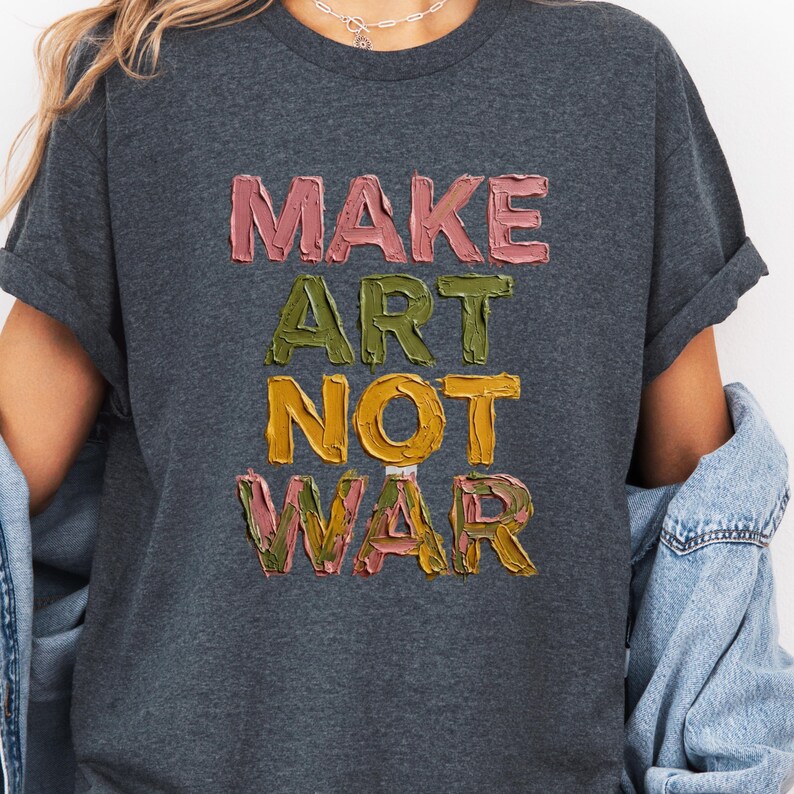 K&ouml;nnte beinhalten: Dunkelgraues T-Shirt mit dem Schriftzug "MAKE ART NOT WAR" in strukturierten, farbenfrohen Buchstaben. Der Text ist in Rosa, Gr&uuml;n, Gelb und Rosa gehalten. Das T-Shirt hat hochgekrempelte &Auml;rmel.