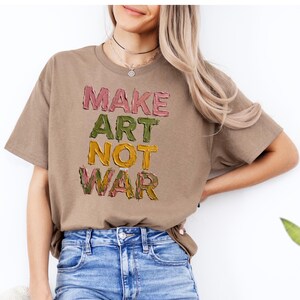 K&ouml;nnte beinhalten: Ein beiges T-Shirt mit dem Schriftzug "MAKE ART NOT WAR" in einer strukturierten, mehrfarbigen Schriftart. Die W&ouml;rter sind vertikal gestapelt, wobei jedes Wort eine andere Farbe hat: rosa, gr&uuml;n, gelb und rosa. Das T-Shirt wird mit einer blauen Jeans getragen.