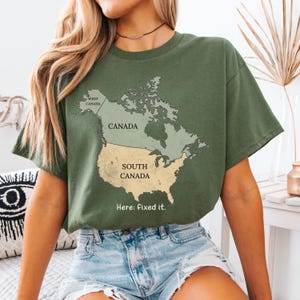 May include: An olive green t-shirt featuring a map graphic of North America. The map labels Canada as Canada and the United States as South Canada, with the text Here: fixed it. printed below the map in a simple, dark-colored sans-serif font.