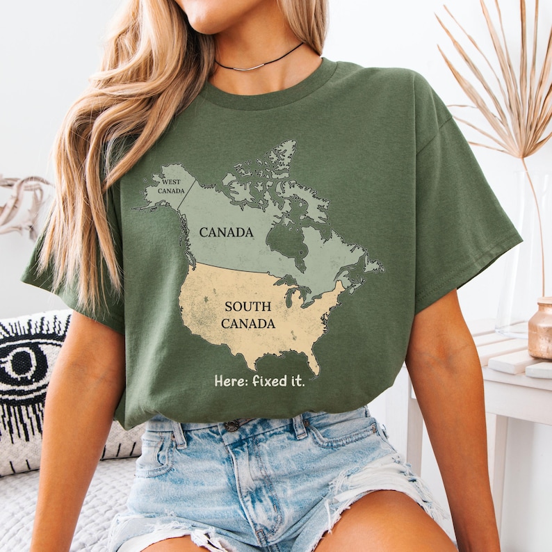 May include: An olive green t-shirt featuring a map graphic of North America. The map labels Canada as Canada and the United States as South Canada, with the text Here: fixed it. printed below the map in a simple, dark-colored sans-serif font.