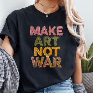 K&ouml;nnte beinhalten: Schwarzes T-Shirt mit dem Schriftzug "MAKE ART NOT WAR" in einem strukturierten, farbenfrohen Schriftzug. Der Text ist vertikal gestapelt, jedes Wort in einer anderen Farbe: Rosa, Gr&uuml;n und Gelb. Ein l&auml;ssiges, k&uuml;nstlerisches Statement.