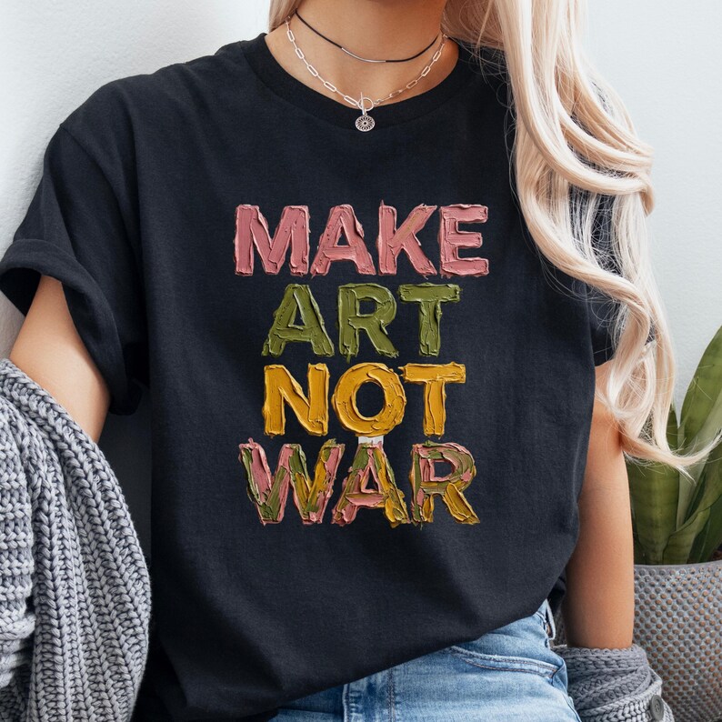 K&ouml;nnte beinhalten: Schwarzes T-Shirt mit dem Schriftzug "MAKE ART NOT WAR" in einem strukturierten, farbenfrohen Schriftzug. Der Text ist vertikal gestapelt, jedes Wort in einer anderen Farbe: Rosa, Gr&uuml;n und Gelb. Ein l&auml;ssiges, k&uuml;nstlerisches Statement.