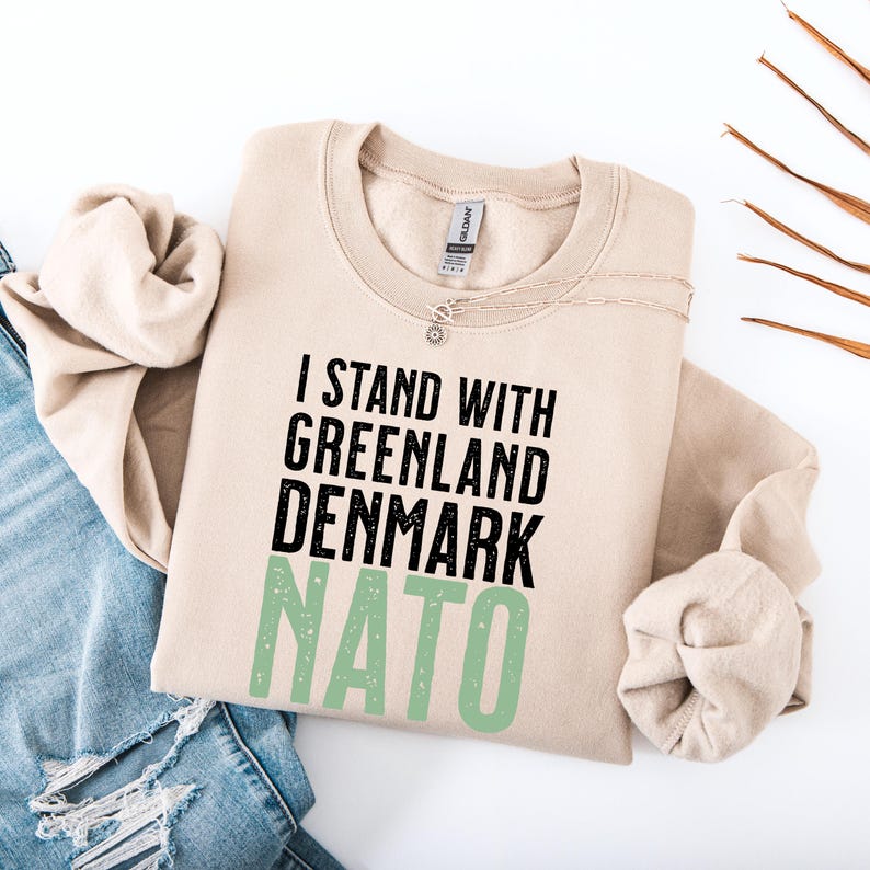 Op de afbeelding: Een beige sweatshirt met de tekst "I STAND WITH GREENLAND DENMARK NATO" in zwart en groen. De sweatshirt is op een blauwe spijkerbroek gevouwen. Een zilveren ketting is zichtbaar.