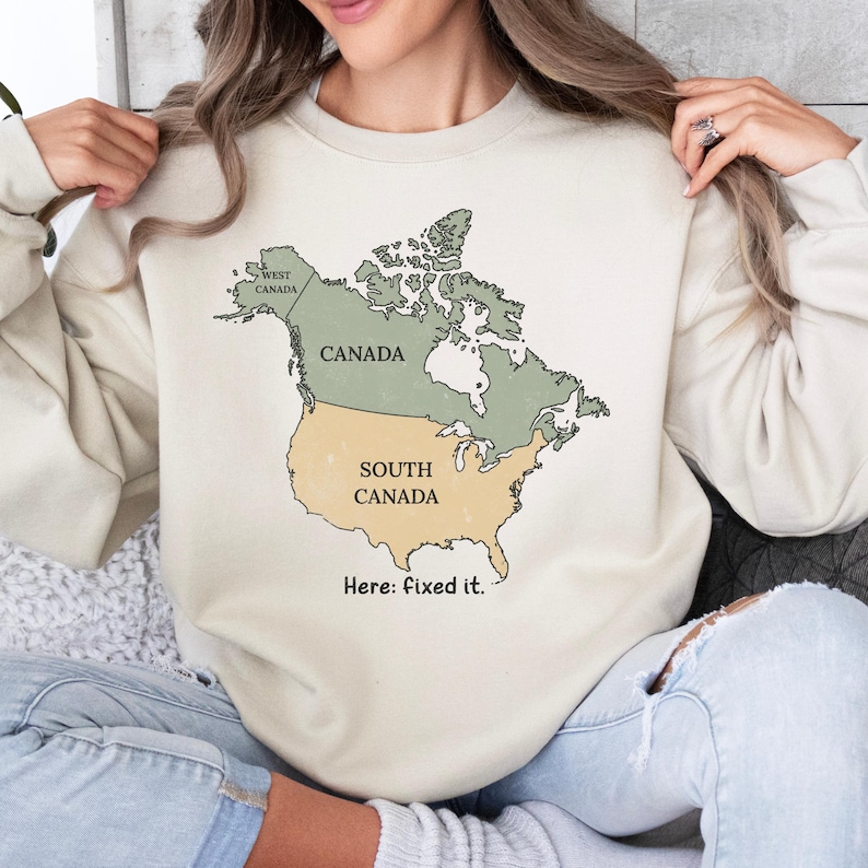 May include: A beige crewneck sweatshirt featuring a graphic map of North America. The map is divided into sections labeled Canada, West Canada, and South Canada. Below the map, the text reads, Here: fixed it. The design uses muted green and tan colors.