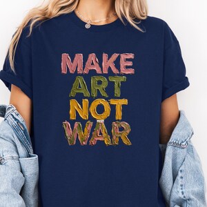 K&ouml;nnte beinhalten: Marineblaues T-Shirt mit dem Text "MAKE ART NOT WAR" in einer strukturierten, farbenfrohen Schriftart. Die Schriftz&uuml;ge sind in Rosa-, Gr&uuml;n-, Gelb- und Braunt&ouml;nen gehalten. Das Shirt hat kurze &Auml;rmel und einen Rundhalsausschnitt.