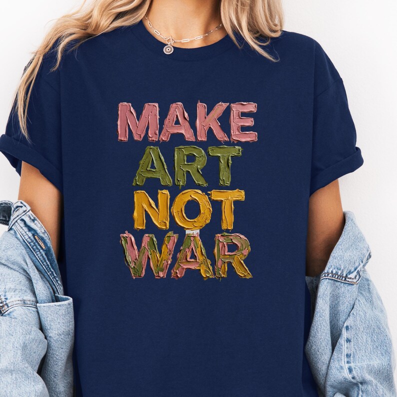 K&ouml;nnte beinhalten: Marineblaues T-Shirt mit dem Text "MAKE ART NOT WAR" in einer strukturierten, farbenfrohen Schriftart. Die Schriftz&uuml;ge sind in Rosa-, Gr&uuml;n-, Gelb- und Braunt&ouml;nen gehalten. Das Shirt hat kurze &Auml;rmel und einen Rundhalsausschnitt.