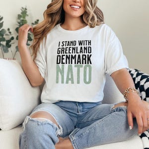 Op de afbeelding: Wit t-shirt met de tekst "I STAND WITH GREENLAND DENMARK NATO" in zwart en groen. Het shirt wordt gedragen door een persoon die een blauwe spijkerbroek draagt.