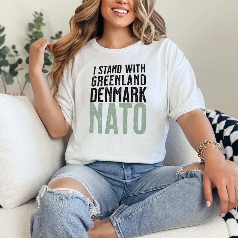 Op de afbeelding: Wit t-shirt met de tekst "I STAND WITH GREENLAND DENMARK NATO" in zwart en groen. Het shirt wordt gedragen door een persoon die een blauwe spijkerbroek draagt.