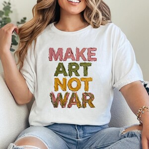 K&ouml;nnte beinhalten: Wei&szlig;es T-Shirt mit dem Text "MAKE ART NOT WAR" in farbenfrohen, strukturierten Buchstaben. Das Shirt hat einen l&auml;ssigen Stil und ist f&uuml;r den Alltag geeignet. Der Text ist in Rosa-, Gr&uuml;n-, Gelb- und Braunt&ouml;nen gehalten.