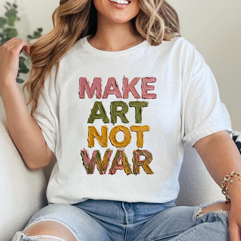 K&ouml;nnte beinhalten: Wei&szlig;es T-Shirt mit dem Text "MAKE ART NOT WAR" in farbenfrohen, strukturierten Buchstaben. Das Shirt hat einen l&auml;ssigen Stil und ist f&uuml;r den Alltag geeignet. Der Text ist in Rosa-, Gr&uuml;n-, Gelb- und Braunt&ouml;nen gehalten.