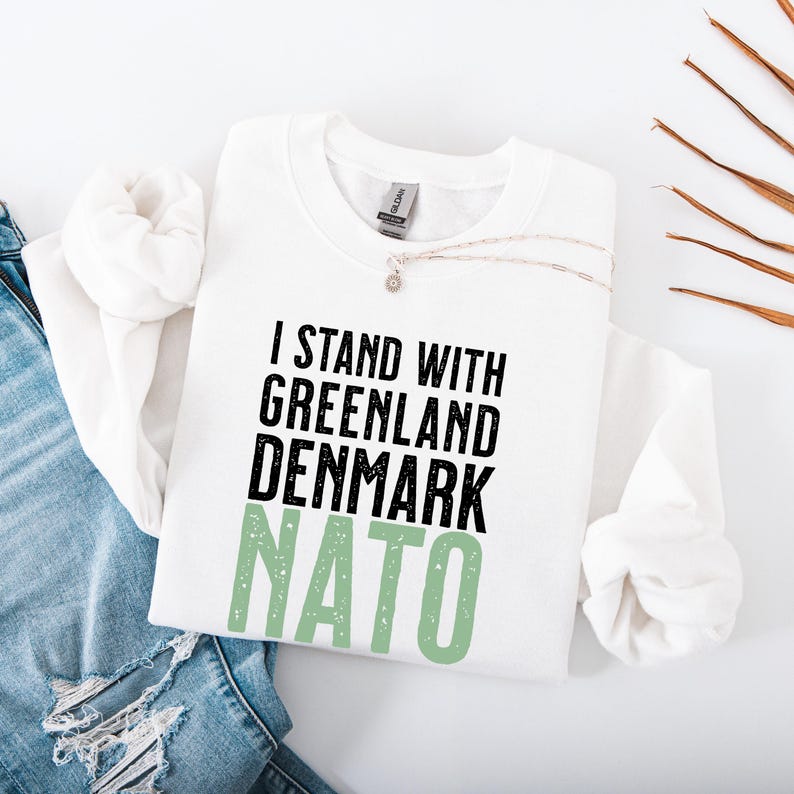 Op de afbeelding: Witte sweatshirt met de tekst "I STAND WITH GREENLAND DENMARK NATO" in zwart en groen. Een zilveren ketting met een kleine hanger ligt op de sweatshirt. De sweatshirt is opgevouwen op een blauwe jeans.