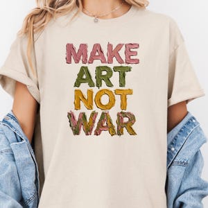 K&ouml;nnte beinhalten: Beiges T-Shirt mit dem Schriftzug "MAKE ART NOT WAR" in farbenfrohen, strukturierten Buchstaben. Der Text ist vertikal angeordnet, wobei jedes Wort eine andere Farbe hat: Rosa, Gr&uuml;n, Gelb und eine Mischung aus Gr&uuml;n und Rosa.