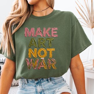 K&ouml;nnte beinhalten: Olivgr&uuml;nes T-Shirt mit dem Text "MAKE ART NOT WAR" in strukturierten, farbenfrohen Buchstaben. Der Text ist in Rosa, Gr&uuml;n, Gelb und Rosa gehalten. Das Shirt hat eine lockere Passform und kurze &Auml;rmel.