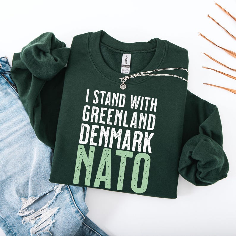 Op de afbeelding: Donkergroene sweatshirt met de tekst "I STAND WITH GREENLAND DENMARK NATO" in wit en lichtgroen. Een zilveren ketting met een ronde hanger ligt op de sweatshirt. De sweatshirt ligt naast een spijkerbroek.