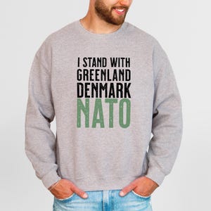 Op de afbeelding: Een lichtgrijs sweatshirt met de tekst "I STAND WITH GREENLAND DENMARK NATO" in zwarte en groene letters. Het sweatshirt is een casual stijl, geschikt voor dagelijks gebruik.