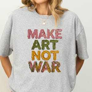K&ouml;nnte beinhalten: Ein hellgraues Sweatshirt mit dem Schriftzug "MAKE ART NOT WAR" in einer farbenfrohen, strukturierten Schriftart. Der Text ist vertikal angeordnet, wobei jedes Wort eine andere Farbe hat: Rosa, Gr&uuml;n und Gelb. Das Sweatshirt hat einen Rundhalsausschnitt und kurze &Auml;rmel.