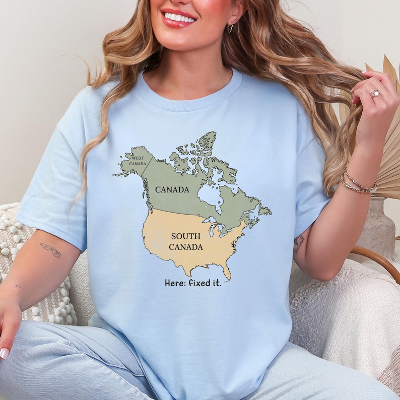 May include: A light blue t-shirt featuring a map of North America with Canada in green and the United States labeled as South Canada in tan. The text West Canada is printed in the northwest region, and the text Here: fixed it. is below the map.