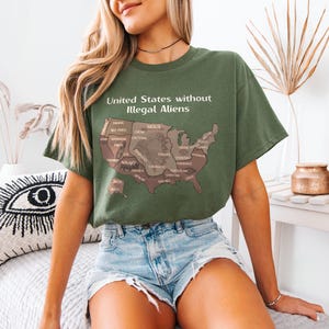 K&ouml;nnte beinhalten: Olivgr&uuml;nes T-Shirt mit dem Text "United States without Illegal Aliens" und einer braunen Karte der USA. Die Karte zeigt die Namen der Bundesstaaten. Das T-Shirt wird mit Jeansshorts getragen.