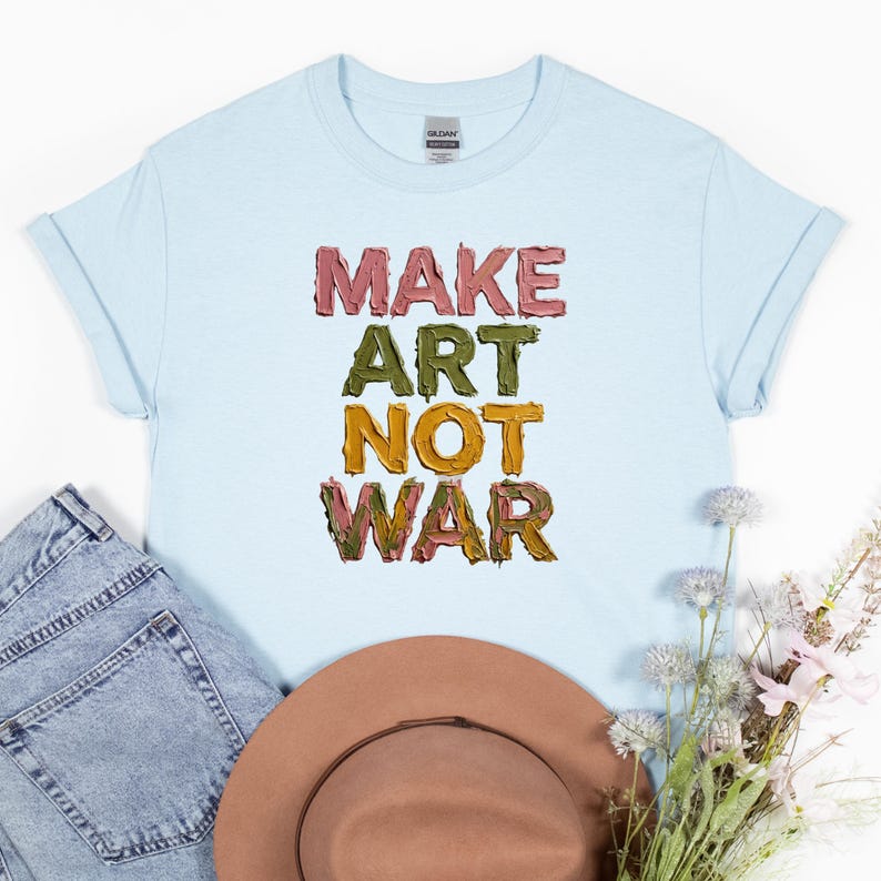 K&ouml;nnte beinhalten: Hellblaues T-Shirt mit dem Text "MAKE ART NOT WAR" in farbenfrohen, strukturierten Buchstaben. Das Shirt wird mit einer Jeans und einem braunen Hut pr&auml;sentiert.