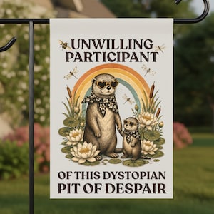 May include: A garden flag featuring an illustration of two otters wearing sunglasses and scarves, standing in front of a rainbow. The flag has the text "UNWILLING PARTICIPANT OF THIS DYSTOPIAN PIT OF DESPAIR."