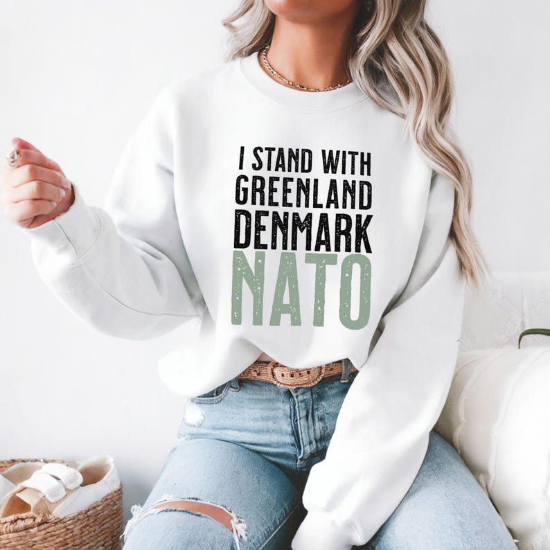 Op de afbeelding: Witte sweatshirt met de tekst "I STAND WITH GREENLAND DENMARK NATO" in zwarte en groene letters. De sweatshirt heeft een ronde hals.