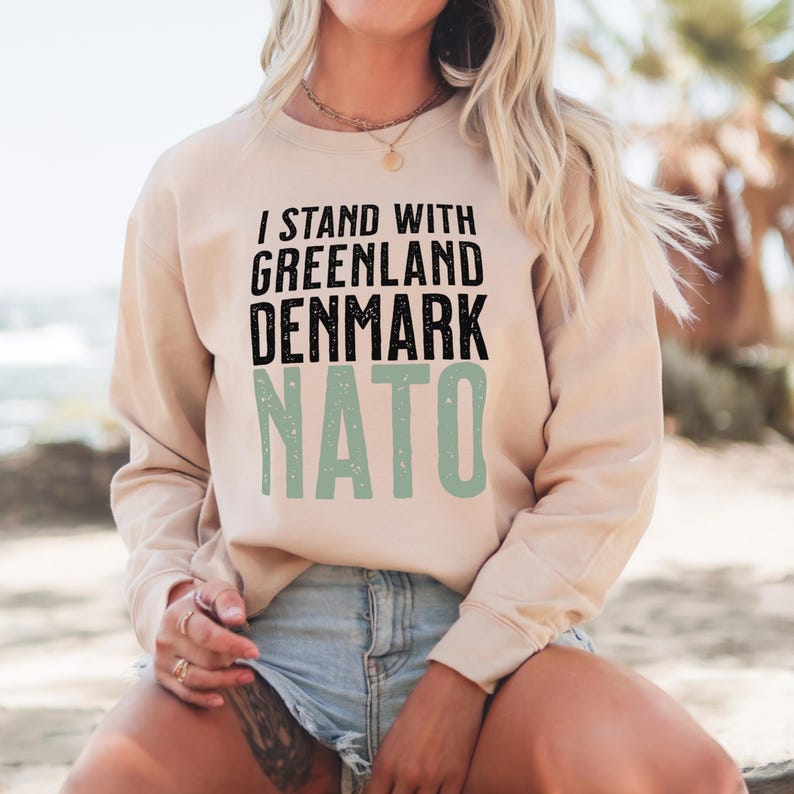 Op de afbeelding: Een beige sweatshirt met de tekst "I STAND WITH GREENLAND DENMARK NATO" in zwart en groen. De lange mouwen sweatshirt heeft een ronde hals en de tekst is op de voorkant gedrukt.