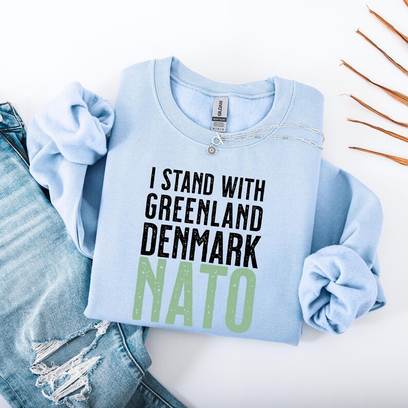 Op de afbeelding: Een lichtblauw sweatshirt met de tekst "I STAND WITH GREENLAND DENMARK NATO" in zwart en groen. Het sweatshirt is opgevouwen met de mouwen naar binnen. Een zilveren ketting ligt bovenop het sweatshirt.