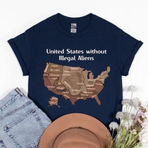 K&ouml;nnte beinhalten: Marineblaues T-Shirt mit dem Text "United States without Illegal Aliens" &uuml;ber einer braunen Karte der Vereinigten Staaten, mit Namen amerikanischer Ureinwohner. Eine blaue Jeans und ein brauner Hut liegen neben dem T-Shirt.