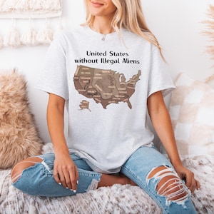 K&ouml;nnte beinhalten: Hellgraues T-Shirt mit einer braunen Karte der Vereinigten Staaten und den Namen der Bundesstaaten. Der Text "United States without Illegal Aliens" ist &uuml;ber der Karte aufgedruckt. Das T-Shirt wird getragen.