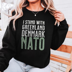 Op de afbeelding: Zwart sweatshirt met de tekst "I STAND WITH GREENLAND DENMARK NATO" in wit en groen. De tekst is in een vetgedrukte, schreefloze lettertype. De sweatshirt is een crew neck stijl en lijkt gemaakt van een zacht materiaal.