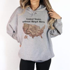K&ouml;nnte beinhalten: Ein hellgrauer Kapuzenpullover mit einer Grafik der Vereinigten Staaten in Braunt&ouml;nen. Der Text "United States without Illegal Aliens" ist &uuml;ber der Karte aufgedruckt. Der Hoodie hat eine Kapuze mit Kordelzug und eine Vordertasche.