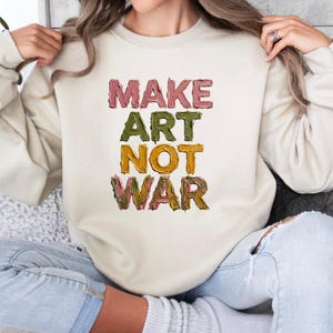 K&ouml;nnte beinhalten: Cremefarbenes Sweatshirt mit dem Text "MAKE ART NOT WAR" in einer farbenfrohen, strukturierten Schrift. Die Buchstaben sind in Rosa-, Gr&uuml;n- und Gelbt&ouml;nen gehalten. Das Sweatshirt ist ein l&auml;ssiger, bequemer Stil.
