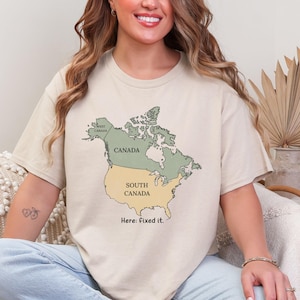 May include: A beige short-sleeve t-shirt featuring a graphic map of North America. The map shows Canada in green and the United States labeled as South Canada in yellow. The text below the map reads, Here: fixed it. The shirt has a crew neckline.