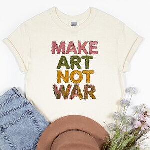 K&ouml;nnte beinhalten: Cremefarbenes T-Shirt mit dem Text "MAKE ART NOT WAR" in einer farbenfrohen, strukturierten Schrift. Das Shirt ist gefaltet, mit einer Jeans und einem braunen Hut im Hintergrund. Ein l&auml;ssiges Kleidungsst&uuml;ck.