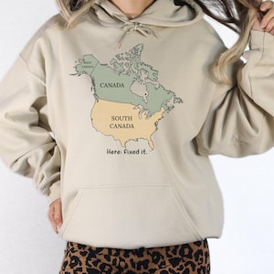May include: A beige hooded sweatshirt featuring a graphic map of North America. The map labels Canada in green and the United States as South Canada in yellow, with the text West Canada and the phrase Here: fixed it. printed below the map.