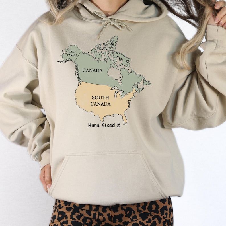 May include: A beige hooded sweatshirt featuring a graphic map of North America. The map labels Canada in green and the United States as South Canada in yellow, with the text West Canada and the phrase Here: fixed it. printed below the map.