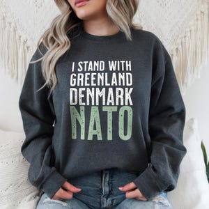 Op de afbeelding: Donkergrijze sweatshirt met de tekst "I STAND WITH GREENLAND DENMARK NATO" in wit en lichtgroen. De crew neck sweatshirt is gemaakt van een zacht materiaal.