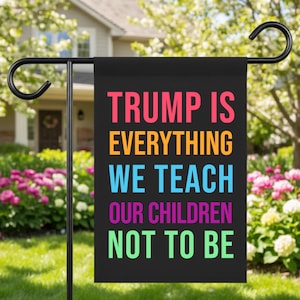 Op de afbeelding: Een zwarte tuin vlag met de tekst "TRUMP IS EVERYTHING WE TEACH OUR CHILDREN NOT TO BE" in diverse heldere kleuren. De vlag hangt aan een zwarte metalen standaard in een tuin.