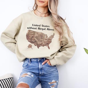 K&ouml;nnte beinhalten: Beiges Sweatshirt mit dem Text "United States without Illegal Aliens" &uuml;ber einer braunen Karte der Vereinigten Staaten. Das Sweatshirt wird mit einer blauen Jeans kombiniert. Das Bild ist von der Taille aufw&auml;rts aufgenommen.