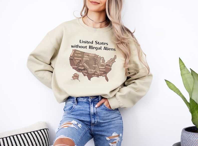 K&ouml;nnte beinhalten: Beiges Sweatshirt mit dem Text "United States without Illegal Aliens" &uuml;ber einer braunen Karte der Vereinigten Staaten. Das Sweatshirt wird mit einer blauen Jeans kombiniert. Das Bild ist von der Taille aufw&auml;rts aufgenommen.