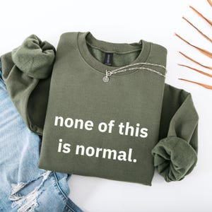 May include: An olive green sweatshirt with the text "none of this is normal" in white. A silver necklace with a pendant is draped on the sweatshirt. The sweatshirt is folded next to a pair of blue jeans.