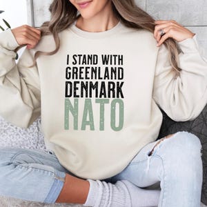 Op de afbeelding: Cr&egrave;mekleurige sweatshirt met de tekst "I STAND WITH GREENLAND DENMARK NATO" in zwarte en groene letters. De sweatshirt is een casual top met lange mouwen.