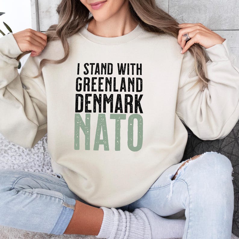Op de afbeelding: Cr&egrave;mekleurige sweatshirt met de tekst "I STAND WITH GREENLAND DENMARK NATO" in zwarte en groene letters. De sweatshirt is een casual top met lange mouwen.
